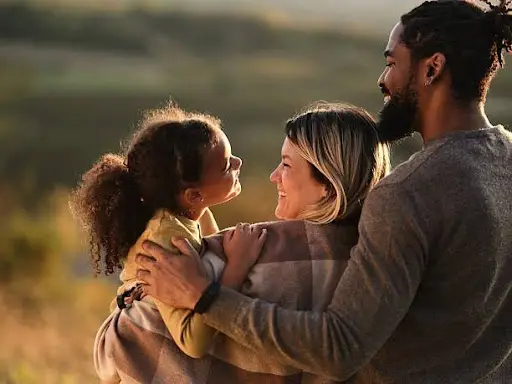 A happy interracial family. A Black father and a white mother holding her mixed-race daughter—smiling together in a grassy field at sunset, representing the internal harmony achieved through IFS therapy.