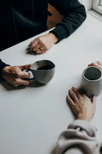 Two people holding coffee mugs while sitting at a table together, illustrating a focused communication check-in recommended by a couples therapist.